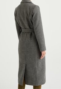 Cappotto lungo con cintura in motivo pied de poule bianco e nero. Presenta una spacco sul retro e un colletto strutturato, realizzato in tessuto strutturato.