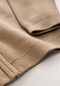 Manica e polsino di un maglione lavorato a maglia beige ripiegati su un tessuto di maglia beige piatto con orlo a coste visibile.