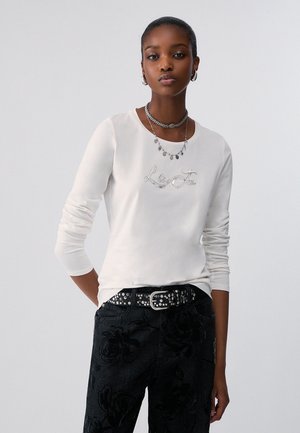 Giovane donna che indossa una camicia bianca a maniche lunghe con testo "Le To", pantaloni neri floreali, cintura borchiata, orecchini a cerchio e collane d’argento sovrapposte.