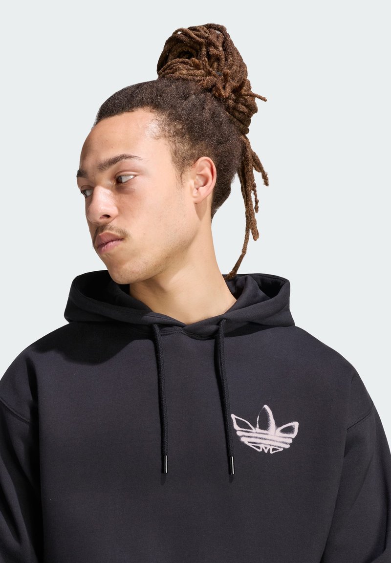 Jeune homme aux dreadlocks attachées porte un sweat à capuche noir avec le logo Adidas blanc, regardant vers sa gauche sur un fond clair uni.