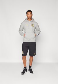 Šedá mikina s kapucí a přední kapsou, zlaté akcenty a logo "MAN UNITED"; spárováno s černými šortkami s podobným značením. Černé tenisky.