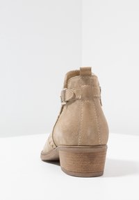 Beige saapas nilkkureissa, joissa on matala pinoheijastin, teksturointi, säädettävä hihna hopeisella soljella ja takan oleva lenkki, joka helpottaa pukemista.