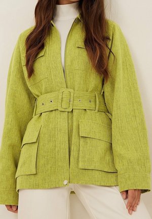 Femme portant une veste ceinturée vert citron avec de grandes poches sur le devant, par-dessus un col roulé blanc et un pantalon blanc cassé, debout sur un fond neutre.