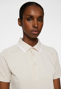 Beige polotröja med vit krage. Har knappstängning och textilstruktur på axlarna. Slätt material, korta ärmar.