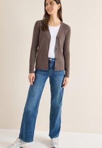 Brązowy prążkowany kardigan z dekoltem w serek, z przodu na guziki, noszony na białej koszuli i zestawiony z niebieskimi szerokimi spodniami dżinsowymi oraz srebrnymi sneakersami.