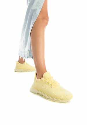 Zapatillas deportivas de malla amarilla con parte superior texturizada, diseño de cordones y suela acolchada. Primer plano del pie y el tobillo, con denim azul claro.