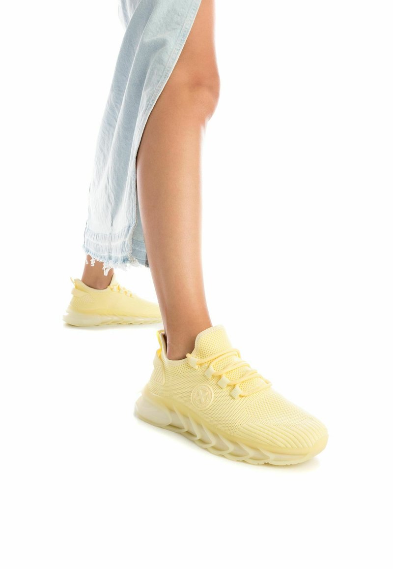 Zapatillas deportivas de malla amarilla con parte superior texturizada, diseño de cordones y suela acolchada. Primer plano del pie y el tobillo, con denim azul claro.