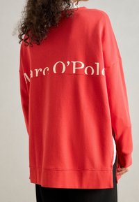 Rode sweatshirt met een ontspannen pasvorm, voorzien van een witte opdruk "Marc O'Polo" over de achterkant en zijsplitten aan de zoom. Gemaakt van zachte, gestructureerde stof.