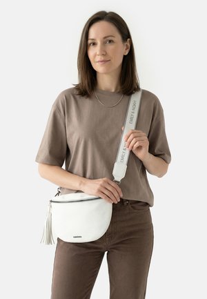 Woman with shoulder-length brown hair wearing a taupe T-shirt and pants, holding a white crossbody bag with a wide strap.