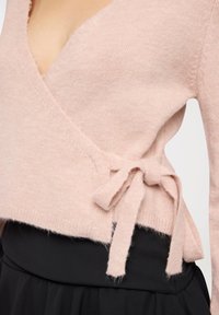 Maglione rosa in misto lana con scollo a V profondo, dettaglio a nodo laterale e finitura testurizzata. Abbinato a una gonna nera, evidenziando uno stile a strati.