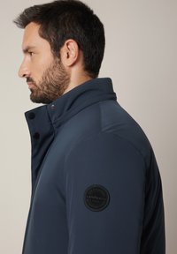 Mężczyzna z ciemną brodą, ubrany w granatową kurtkę z wysokim kołnierzem, zwrócony w lewo, z widoczną okrągłą czarną łatą na rękawie.