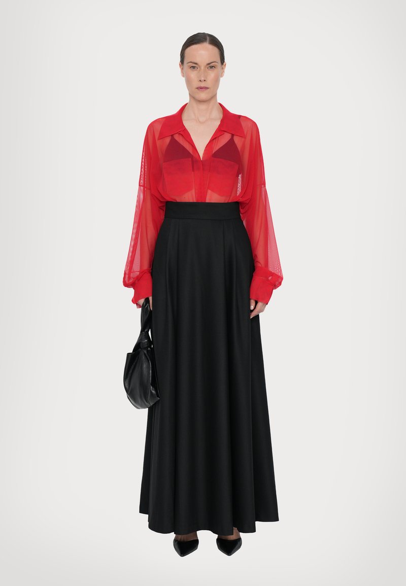 Blusa rossa trasparente con maniche lunghe, abbinata a una gonna maxi nera. Gli accessori includono una borsa nera e scarpe nere a punta.