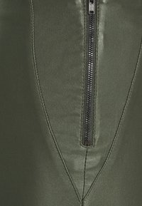 Olivgrünes Kunstleder mit einer glatten Textur. Verfügt über einen zentralen Reißverschluss, umrahmt von schrägen Nähten.