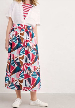 Woman wearing a colorful geometric patterned skirt, red and white striped top, white short-sleeve shirt, and white sneakers standing against a plain wall.