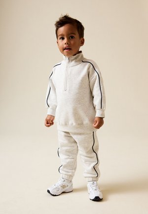Unge barn iført en lysegrå træningsdragt med sorte kantninger og hvide sneakers står mod en ensfarvet beige baggrund.