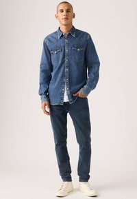 Džinsinis marškinis su klasikinio kirpimo, dviem krūtine kišenėmis, šviesiomis siūlėmis ir vidutinio mėlynumo spalva, derinamas su tamsiais džinsais ir baltomis sportinėmis batelėmis.