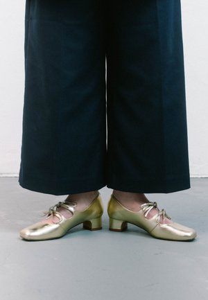 Scarpe in pelle dorata con punta quadrata e piccolo tacco largo, caratterizzate da un design a ritaglio rotondo e dettagli legati sulla parte anteriore.