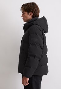 Schwarze Puffy-Jacke mit Kapuze, Reißverschluss vorne und seitlichen Taschen. Hat eine strukturierte Oberfläche und ein Logo-Patch am Ärmel.