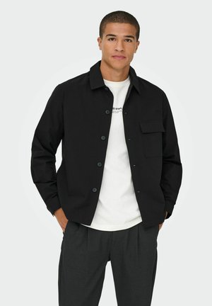 Veste noire à col, à boutons, avec une poche poitrine simple et une texture lisse, portée sur un t-shirt blanc et un pantalon gris.