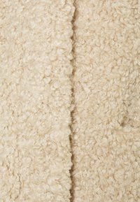 Beiger Stoff mit strukturierter Oberfläche und flauschiger, lockiger Textur, der ein weiches und warmes Aussehen aufweist, typisch für Schaffell oder Kunstschaffell.