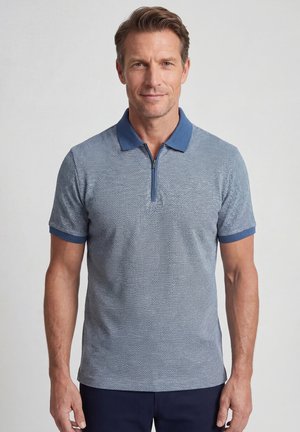 Man draagt een blauw polohemd met korte mouwen en een ritskraag, staande tegen een effen lichte achtergrond.