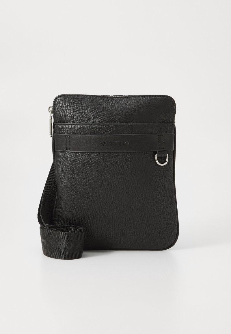 Sac à bandoulière en cuir noir avec une texture lisse, fermeture par zip, sangle avant portant le nom de la marque et sangle en tissu ajustable.