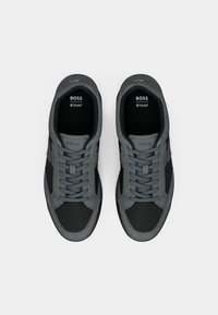 Chaussures de sport grises avec une combinaison de matériaux lisses et texturés. Elles présentent un bout arrondi, un design à lacets et des accents sombres contrastants.