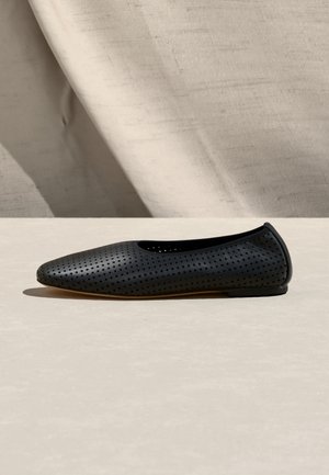 Zapato plano negro perforado con puntera redondeada y tacón bajo sobre una superficie beige con un fondo de tela texturizada.