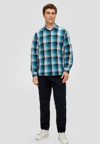 Jonge man in een blauw en zwart geruit overhemd met knopen, zwarte broek en witte sneakers, staande tegen een egale achtergrond.