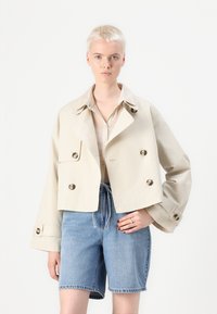 Giacca corta doppiopetto beige con grandi bottoni, maniche lunghe e colletto testurizzato, abbinata a pantaloni shorts in denim chiaro.