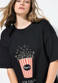 Camiseta negra de gran tamaño con un diseño gráfico de un cubo de palomitas de maíz con rayas rojas y blancas, adornado con pequeñas perlas blancas y texto.