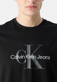 Camiseta de algodón negra con cuello acanalado que presenta un gran gráfico gris de "CK" y el texto "Calvin Klein Jeans" en blanco debajo.