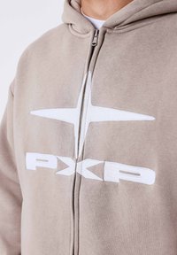 Beige hoodie met rits, gemaakt van zacht materiaal, met een wit logo en ontwerp op de voorkant, een gladde textuur en een standaard pasvorm.