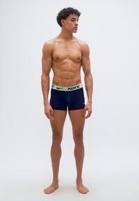 Νέος άνδρας με σγουρά μαλλιά που φοράει μπλε ναυτικό εσώρουχο Nike boxer, στέκεται ξυπόλυτος μπροστά σε απλό λευκό φόντο, κοιτάζοντας προς το πλάι.