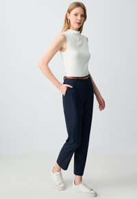 Witte mouwloze top met een hoge hals, gecombineerd met marineblauwe tapershorts en witte sneakers. Bruine riem met een ronde gouden gesp als accent.