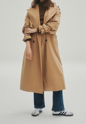 Person iført en lang beige trenchcoat, mørkegrøn top, blå jeans og hvide sneakers med sorte striber på en ensartet lys baggrund.