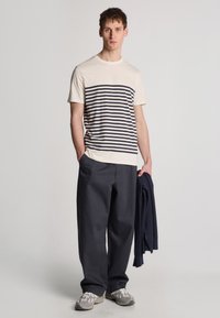 Gestreepte t-shirt in crème en marine, van katoenmix. Gepaard met losse marineblauwe broeken en grijze sneakers. Model houdt een marineblauwe trui vast.