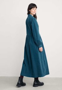 Abito lungo in corduroy color teal con vita arricciata e maniche a sbuffo, abbinato a stivali neri alla caviglia. Texture morbida e silhouette fluida.