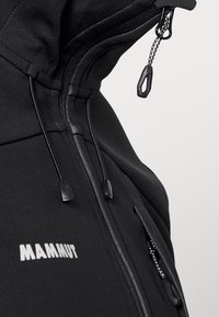 Giacca softshell nera con zip anteriore, cappuccio regolabile, tessuto testurizzato e logo bianco "MAMMUT" sul petto. Dettagli hardware visibili.