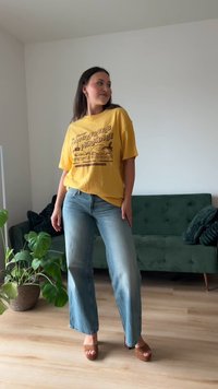 Γυναίκα που στέκεται μέσα σε κλειστό χώρο, φοράει κίτρινη μπλούζα με σχέδιο, ανοιχτόχρωμα μπλουτζίν και καφέ σανδάλια με τακούνι, με πράσινο καναπέ και φυτά στο φόντο.