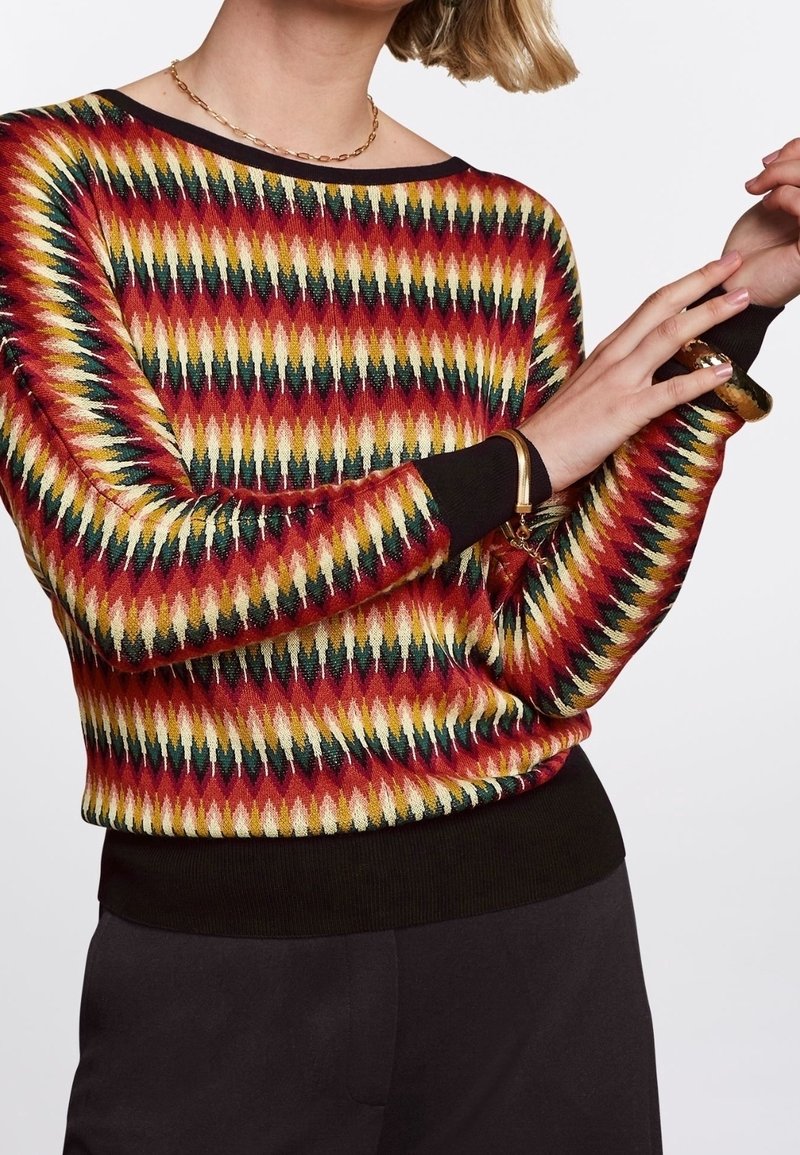 Femme portant un pull à motif zigzag multicolore avec des poignets et un ourlet noirs, ajustant un bracelet à son poignet.
