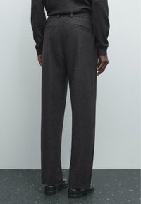Pantalons en laine gris foncé avec une coupe droite, taille élastique et deux poches arrière, associés à des chaussures noires sur une surface en pierre.