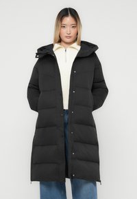 En ung kvinde iført en lang sort puffjakke med hætte over en hvid zip-up sweater og blåjeans, står op ad en ensfarvet hvid baggrund.