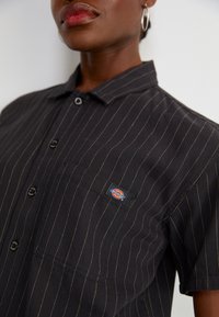 Svart pinstripe kortermet skjorte med knappelukking, venstre brystlomme, og en synlig merkepute i blå, rød og gul farge.