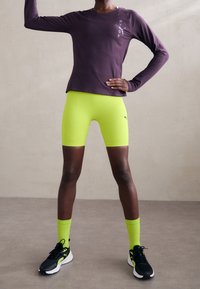 Fioletowa długorękawowa koszulka sportowa, neonowo zielone szorty kompresyjne, czarne sneakersy z białymi podeszwami i neonowo zielone skarpetki. Prosty design i żywe kolory.