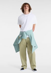 Witte T-shirt, lichtgroene jas om de taille geknoopt, kaki broek en multicolor sneakers. Krullend haar, met het gezicht naar voren.