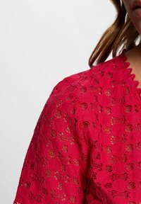 Article en dentelle rouge présentant un col à bord festonné et des motifs circulaires. Le tissu est texturé avec un design délicat à tissage ouvert.