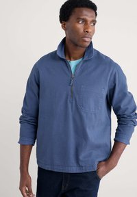 Blå tröja i mjukt material med kvarterdragkedja, bröstficka och långa ärmar. Har en avslappnad passform och synliga sömdetaljer.