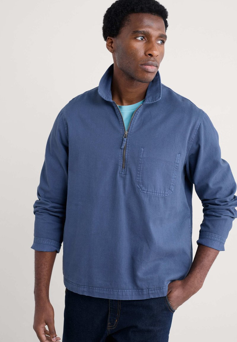 Blå tröja i mjukt material med kvarterdragkedja, bröstficka och långa ärmar. Har en avslappnad passform och synliga sömdetaljer.