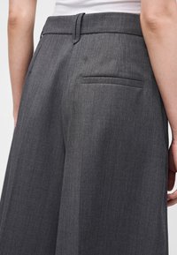 Gros plan d'une personne portant un pantalon gris taille haute avec une poche passepoilée au dos et un haut blanc rentré.